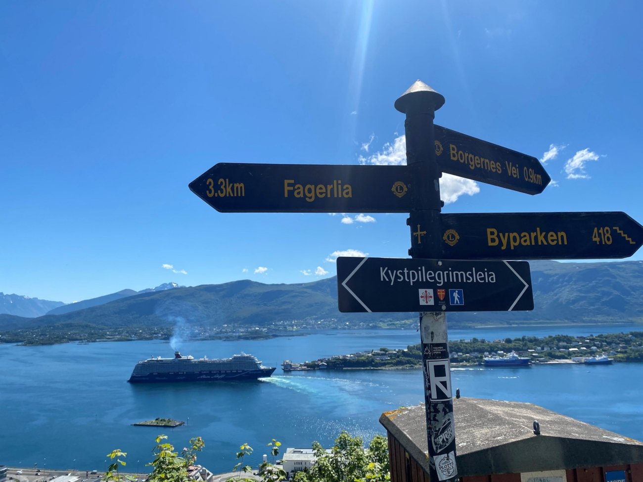 Bilde av Ålesund i solskinn, sett fra byfjellet Aksla. Cruiseskip på fjorden og et skilt som viser vei til ulike destinasjoner,.
