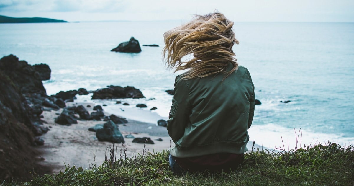 Kvinne sitter på en klippe med utsikt over havet. Vinden tar tak i håret hennes.