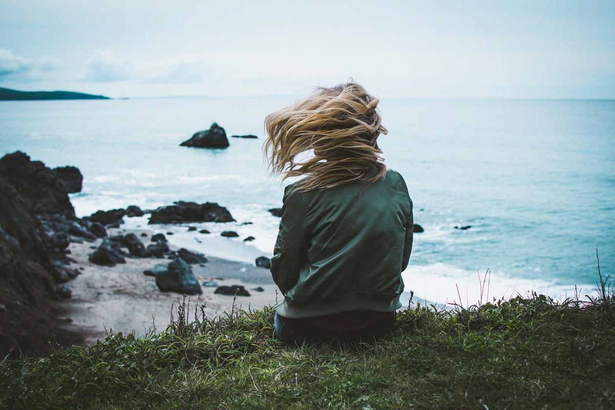 Kvinne sitter på en klippe med utsikt over havet. Vinden tar tak i håret hennes.